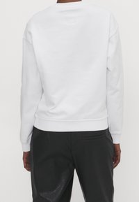 Sudadera blanca con cuello redondo, hombros caídos y puños y dobladillo acanalados. Textura de tejido lisa, diseño sencillo y sin gráficos visibles.