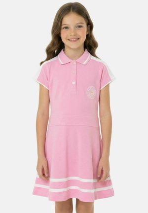 Fille souriante aux cheveux ondulés portant une robe polo rose avec des rayures blanches sur les manches et la jupe, debout devant un fond uni.