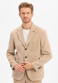 Beige Corduroy-Blazer mit vertikalen Streifen, Reverskragen, zwei Fronttaschen und zwei schwarzen Knöpfen. Getragen über einem weißen Hemd.