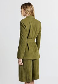 Donna con capelli biondi che indossa un completo verde oliva strutturato, con giacca con cintura e pantaloni larghi a culotte, girata di spalle su uno sfondo bianco.