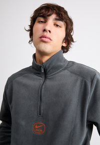 Γκρι fleece polo με φερμουάρ μέχρι το στήθος, που διαθέτει ένα εντυπωσιακό πορτοκαλί λογότυπο Nike AIR στο στήθος και υφή υφάσματος.