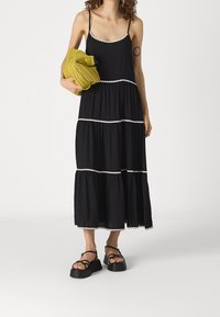 Robe maxi noire avec design à volants, accents en blanc, fines bretelles et silhouette fluide. Le modèle tient un châle texturé vert. Sandales noires à plateforme.