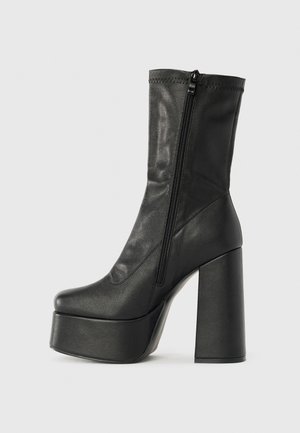 Botín de cuero negro hasta el tobillo con un alto tacón ancho, suela de plataforma y cierre con cremallera lateral. Textura suave con detalle de costura superior.