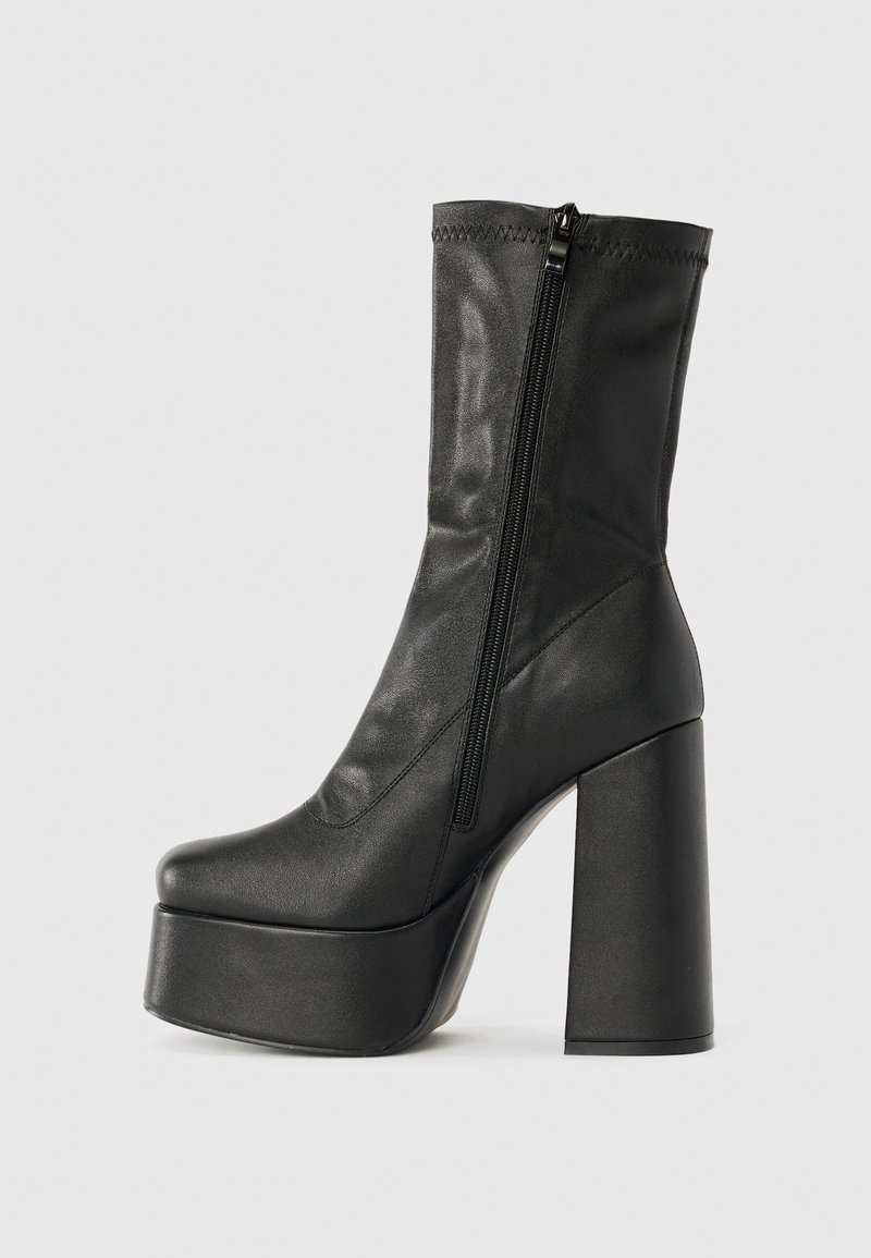 Bota de tornozelo em couro preto com um alto salto bloco, sola com plataforma e um fecho lateral em zíper. Textura suave com detalhes de costura no topo.