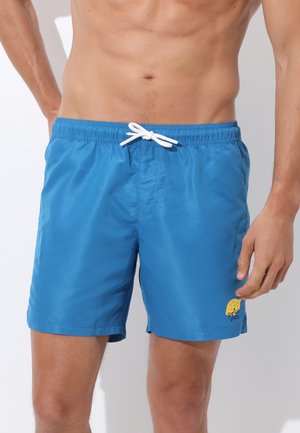 Torace maschile con pantaloncini da bagno blu con coulisse bianca e piccolo logo giallo sulla gamba sinistra, in piedi su sfondo bianco.