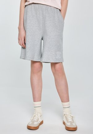 Osoba ubrana w jasno szare krótkie spodenki za kolano z elastycznym pasem, białe skarpety do kostek w paski oraz beżowe trampki, stojąca z rękami w kieszeniach.