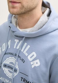 Hanorac cu glugă de culoare albastru deschis, cu interior gri, având text imprimat alb și un logo circular. Textură moale, design casual, șnur ajustabil.