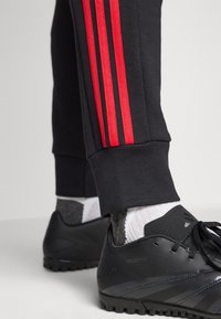 As calças de treino pretas apresentam três riscas vermelhas nas laterais. Os sapatos de futebol pretos com design texturizado e atacadores completam o visual.