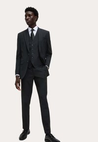 Czarny garnitur trzyczęściowy z dopasowaną marynarką, kamizelką i wąskimi spodniami. Biała koszula z kołnierzykiem i ciemny krawat, zestawione z czarnymi butami wizytowymi.