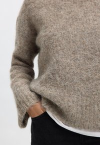 Világosbarna pulóver puha, bolyhos anyagból, laza szabással. Hosszú ujjakkal és széles nyakkivágással rendelkezik, fehér felsővel rétegezve.