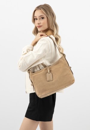 Jeune femme aux longs cheveux blonds portant une veste blanche et une jupe noire, portant un grand sac à bandoulière en suède beige avec une étiquette.