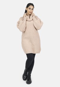 Vestido de punto beige claro con cuello alto, textura acanalada y mangas oversize, combinado con leggings negros y botas con cordones.