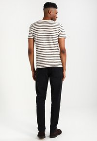 Gestreept beige en marineblauwe T-shirt met korte mouwen, gecombineerd met zwarte jeans. Model staat met de rug naar de camera, waardoor de horizontale lijnen van het shirt zichtbaar zijn.