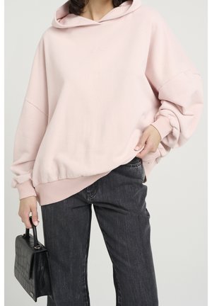 Person iført en løs, lyserød hættetrøje og mørkegrå jeans, der holder en sort tekstureret håndtaske i venstre hånd.