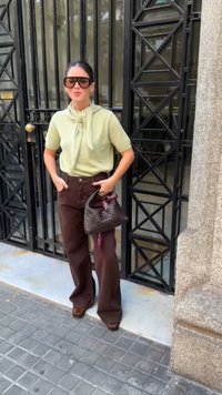 Bluză scurtă de culoare verde deschis cu detalii legate, pantaloni maro cu talie înaltă, ochelari de soare negri oversized și o geantă țesută neagră cu un accent roz.