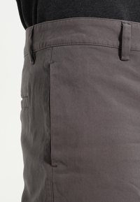 Gros plan sur la taille et la zone des poches d'un pantalon gris porté avec une chemise foncée, montrant les passants de ceinture et la texture du tissu.