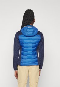 Casaco puffer azul com enchimento horizontal, capuz azul e painéis laterais azul-marinho. O material parece leve e resistente à água.