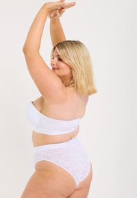 Vitt bandeau-bh med texturerad spetskant och justerbar bakstängning, i kombination med vita högmidjade underkläder med spetsmönster.