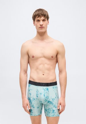 Jeune homme aux cheveux bruns courts portant un caleçon boxer bleu clair à motifs avec une ceinture noire de marque, debout devant un fond clair uni.