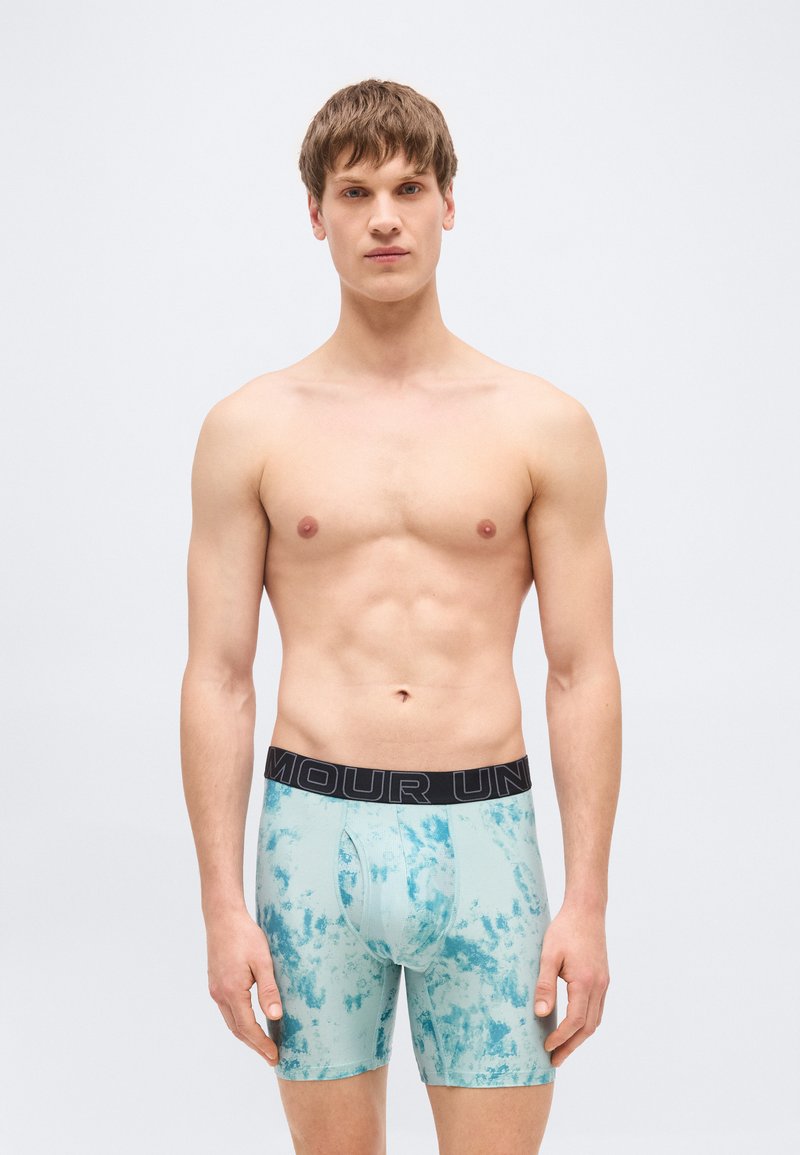 Νέος άνδρας με κοντά καστανά μαλλιά φοράει ανοιχτό μπλε μποξεράκια με σχέδιο και μαύρη ζώνη μέσης με λογότυπο, στέκεται σε απλό φωτεινό φόντο.