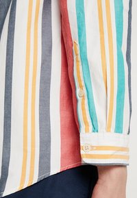 Camicia a righe con linee orizzontali in rosso, blu, bianco e giallo. Presenta un polsino con bottone e una texture di cotone liscia.