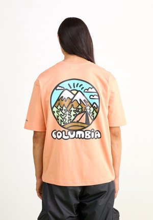 Person with long dark hair wearing a peach shirt featuring a colorful mountain, trees, tent graphic and "Columbia" text on the back.