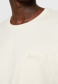 T-shirt à manches courtes couleur écrue en coton, avec une poche arborant un logo "levi's" en relief, col rond et texture douce.