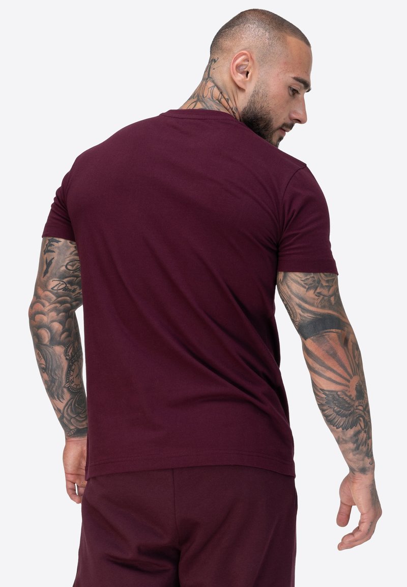 Burgundské tričko s krátkým rukávem, kulatým výstřihem, vyrobené z měkké látky, v přiléhavém střihu, které ukazuje složité tetování na pažích.