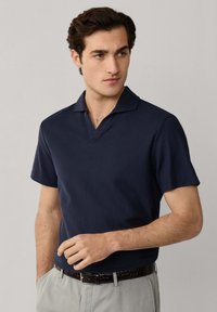 Giovane uomo con capelli scuri che indossa una polo blu navy a maniche corte e pantaloni grigio chiaro, in piedi davanti a uno sfondo chiaro e uniforme.