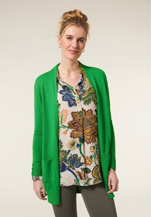 Groene gebreide cardigan met een open voorkant en lange mouwen, gedragen over een bloempatroon blouse. Opmerkelijke zakken en knoopmanchetten.