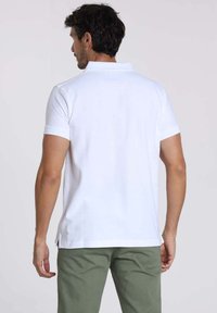 T-shirt polo blanc avec un col classique, des manches courtes et des fentes latérales, porté avec un pantalon vert. La vue de dos montre une texture lisse et des lignes épurées.