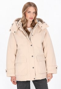 Beige gewatteerde jas met capuchon, drukknoopsluiting, voorzakken en een zachte textuur. Heeft een grijze binnenvoering.