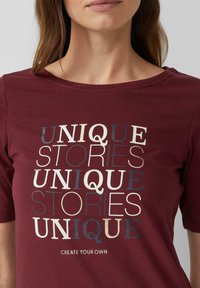 Burgunderrotes T-Shirt mit einem schichtigen Textdesign, das die Wörter "UNIQUE" und "STORIES" in verschiedenen Farben und Größen zeigt. Aus Baumwollmaterial, kurze Ärmel.