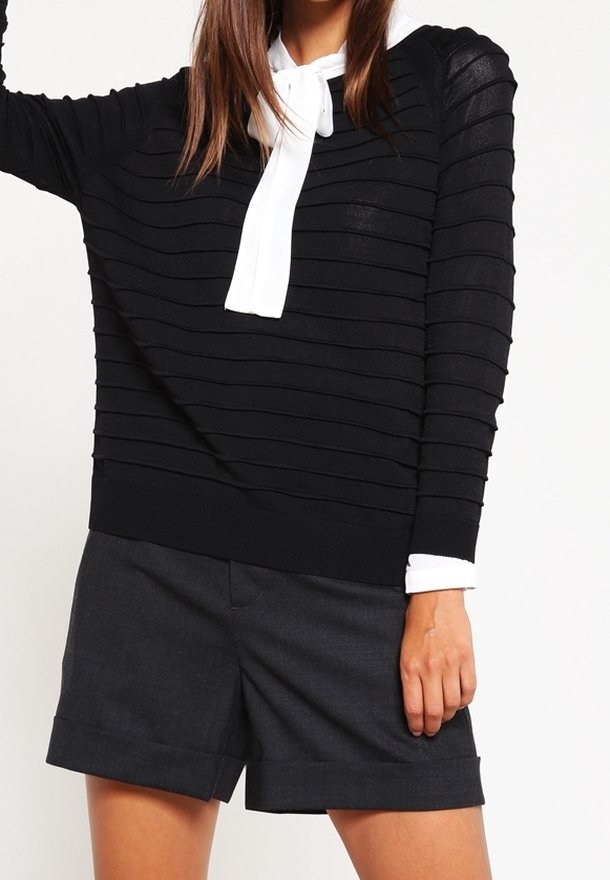 Femme portant un pull noir côtelé avec épaules rembourrées, une écharpe blanche et un short tailleur foncé, debout devant un fond blanc.