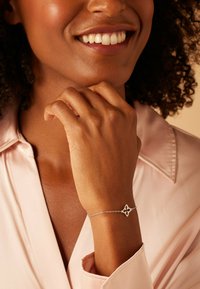 Bracelet en argent avec un design floral, doté d'une fine chaîne et d'un ornement central. Porté sur une main reposant contre le menton.