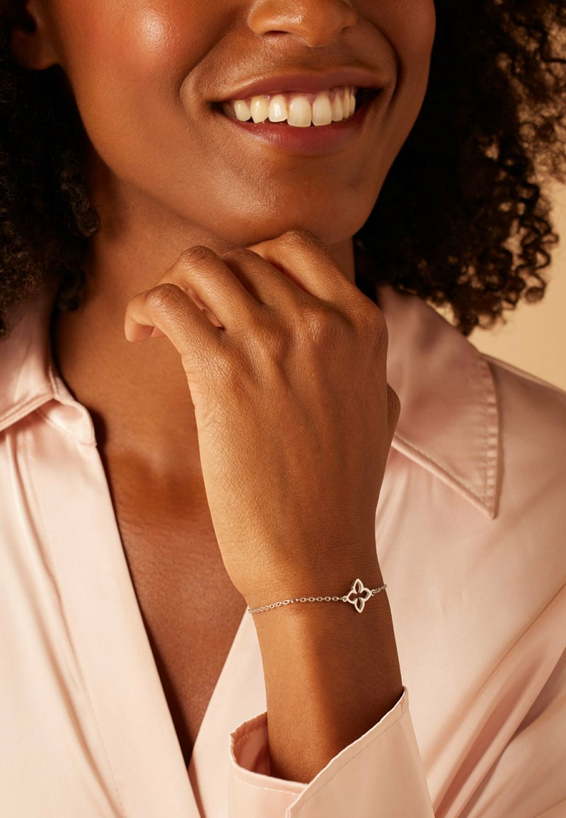 Bracelet en argent avec un design floral, doté d'une fine chaîne et d'un ornement central. Porté sur une main reposant contre le menton.