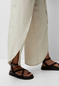 Des sandales plates noires avec des brides croisées et une boucle à la cheville, assorties à un pantalon léger et ample avec des fentes sur les côtés.