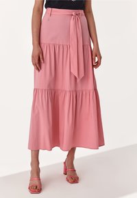 Vrouw in een roze tiered midi-jurf met een gestrikte riem en roze hoge hakken, staand tegen een plain witte achtergrond.