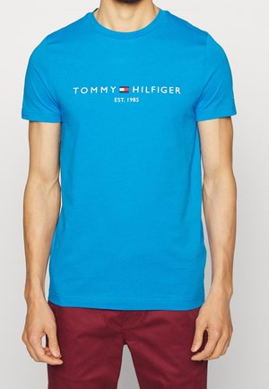 Mand iført en knaldblå Tommy Hilfiger T-shirt med logo og teksten "EST. 1985", kombineret med mørkerøde bukser, mod en ensfarvet baggrund.