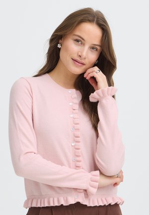Frau mit langen braunen Haaren, die eine rosa gerüschte, knöpfbare Strickjacke, silberne Ohrringe und silberne Ringe trägt und mit der Hand nahe am Gesicht vor weißem Hintergrund posiert.