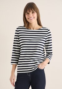 Femme souriante aux cheveux bruns, portant un t-shirt à manches longues rayé noir et blanc et un jean foncé, main dans la poche, fond beige.