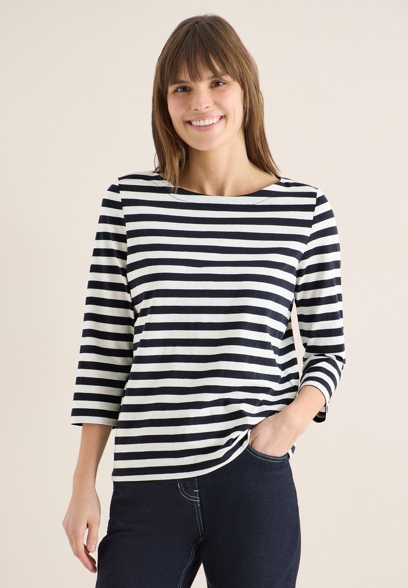 Femme souriante aux cheveux bruns, portant un t-shirt à manches longues rayé noir et blanc et un jean foncé, main dans la poche, fond beige.