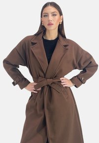 Bruine oversized jas van een gestructureerde stof, met lange mouwen, grote revers, een ceintuur in de taille en zijzakken. Knopmanchetten zorgen voor extra detail.