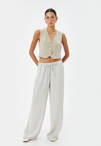 Gilet beige cropped avec un décolleté en V et cinq boutons, associé à un pantalon léger gris clair à coupe ample avec une taille élastique et des liens.