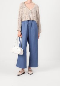 Virkat beige kardiagan med knytfram, tillsammans med lösa blå byxor. Håller en vit handväska; bär ljusbeige platta skor.