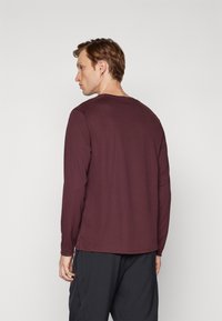 Bordó hosszú ujjú póló kerek nyakkivágással és oldalsó nyílásokkal, puha anyagból készült. Sötét nadrággal viselve, hátulnézetből.