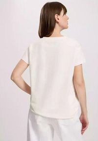 Biała koszulka krótki rękaw z okrągłym dekoltem i gładką fakturą, widoczna z tyłu, zestawiona z jasnymi spodniami.