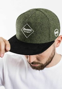 Green herringbone-patterned cap with a black suede brim and white logo patch. Features a classic snapback design with an adjustable strap.