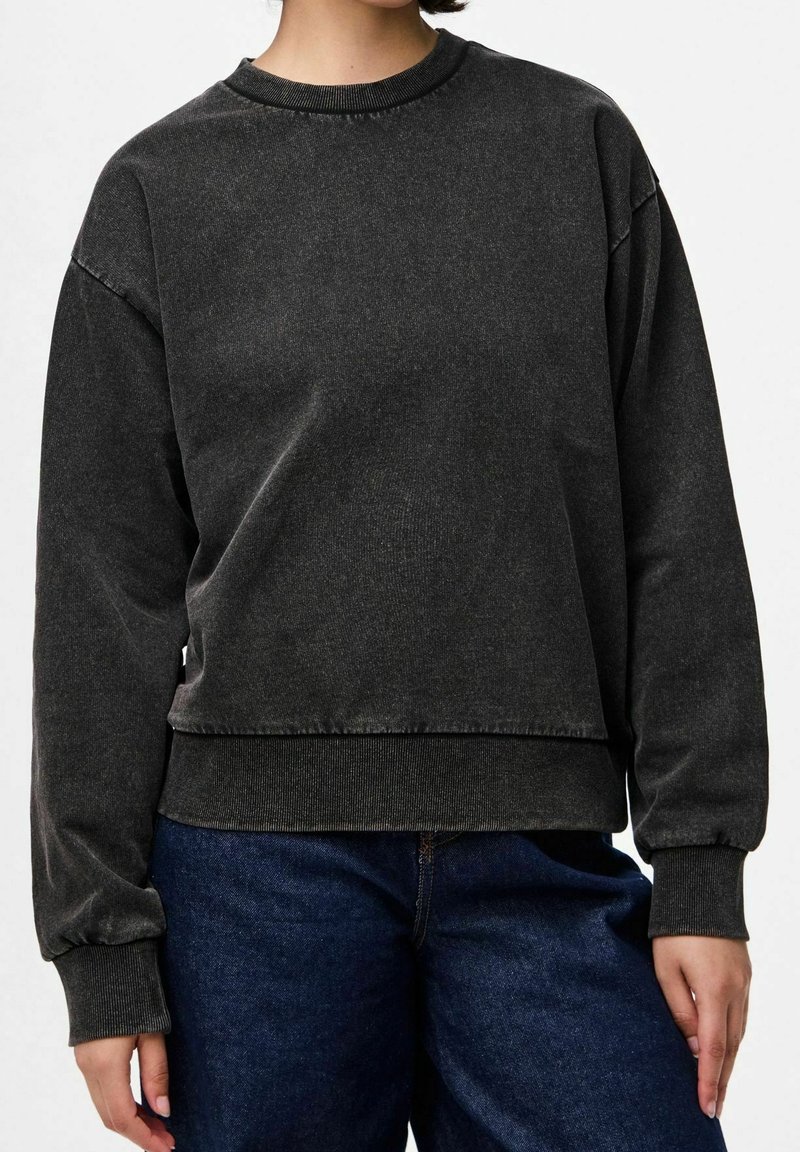 Persoon draagt een gewassen zwarte crewneck trui met geribbelde manchetten en zoom, gecombineerd met donkerblauwe spijkerbroek.