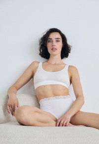 White cotton bralette and high-waisted panties with lace trim. Soft, smooth texture. Model seated against a light background.